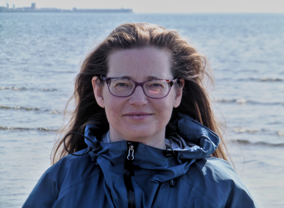 Headshot of Catherine Brys wearing a blue walking jacket with the sea in the background, image credit Catherine Brys