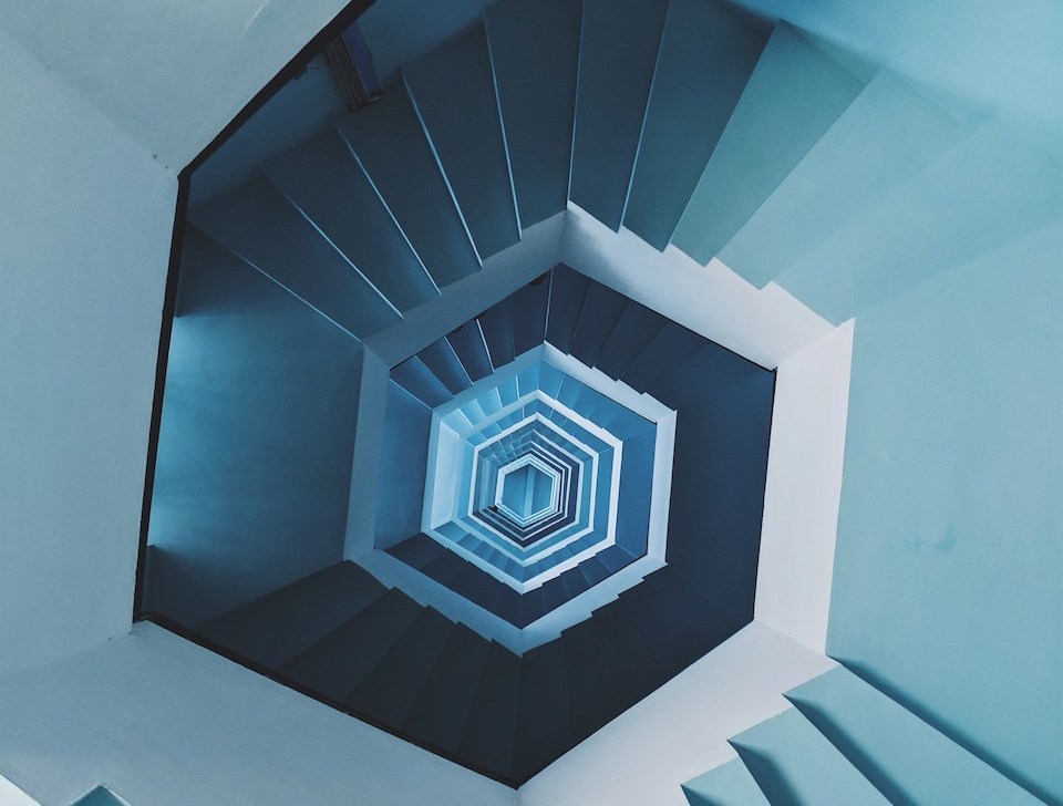 Photo taken with strong perspective by looking down from the top of a very high pastel blue spiral staircase, photo by Faris Mohammed