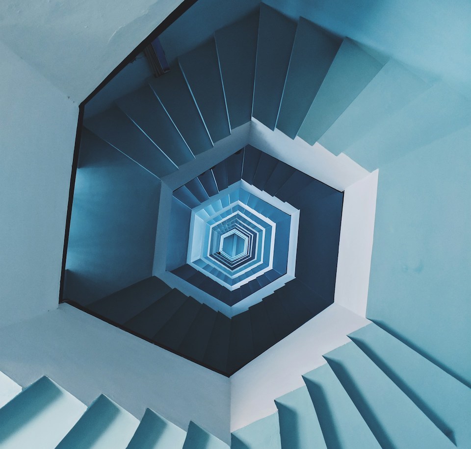 Photo taken with strong perspective by looking down from the top of a very high pastel blue spiral staircase, photo by Faris Mohammed